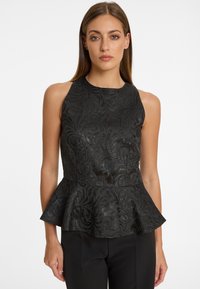 Femme portant un haut peplum noir sans manches avec un motif floral subtil, associé à un pantalon noir, sur un fond blanc uni.