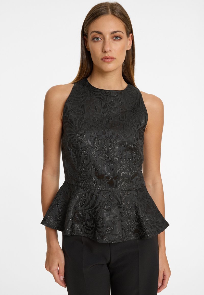 Femme portant un haut peplum noir sans manches avec un motif floral subtil, associé à un pantalon noir, sur un fond blanc uni.