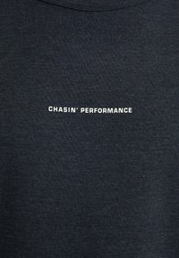 Tummanharmaa puuvillapaita, jossa on uritettu pinta ja rinnassa valkoiset tekstit "CHASIN' PERFORMANCE."