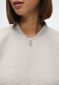 Lys grå bomberjakke med en myk, teksturert fleece-finish. Har en ribbestrikket krage og en sølvfarget glidelås foran.