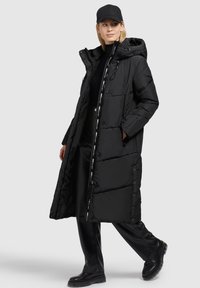 Zwarte gewatteerde jas met een capuchon, lange lengte, frontknopen en een rits. Gekleed met zwarte leren broek en laarzen.