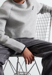 Grå sweatshirt med ribbad halsringning och svart Nike-logotyp, parat med mörka jeans. Modell sitter på en trådstol och visar en avslappnad pose.