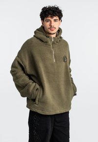 Sudadera de felpa color verde oliva con media cremallera, bolsillo frontal y parche con logo. Tejido texturizado, corte relajado y cordón ajustable en la capucha.