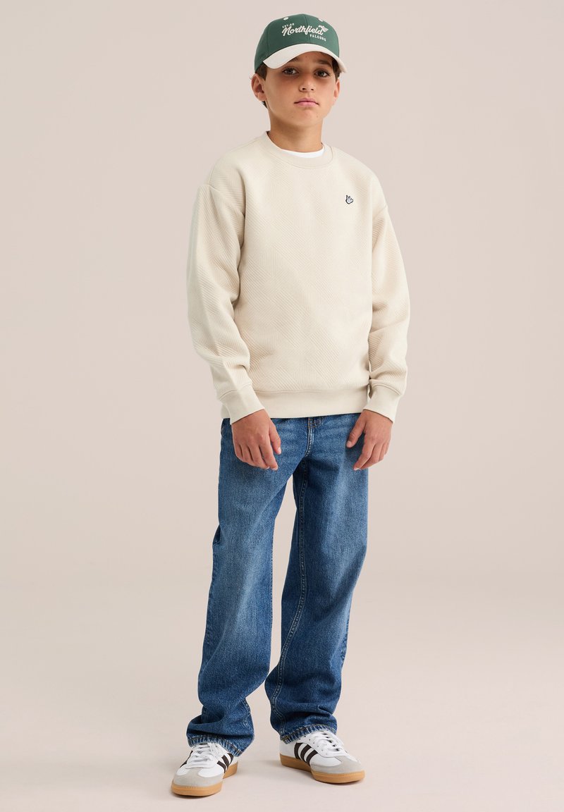 Garçon portant un sweat-shirt crème, un jean bleu, des baskets blanches et une casquette verte, debout devant un fond beige uni.