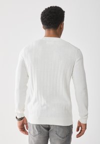 Pull en maille côtelée blanc avec des manches longues, présentant un col rond et une taille ajustée, assorti à un jean gris.