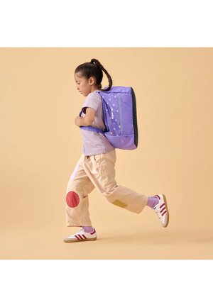 Lila Rucksack mit Punkten, gepolstertes Design, seitlicher Reißverschluss, getragen von einem Kind in beigen Hosen mit bunten Patches und weißen Turnschuhen.