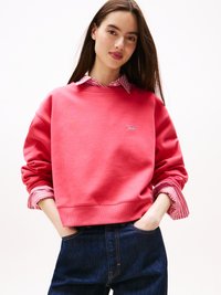 Felpa rossa con collo tondo e maniche lunghe, sovrapposta a una camicia a righe bianche e rosa. Jeans in denim scuro dal taglio classico.