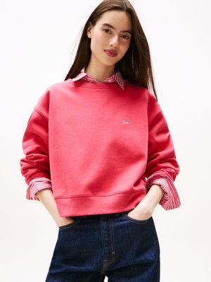 Sweatshirt rouge avec un col ras du cou et des manches longues, superposé sur une chemise rayée blanche et rose. Jean en denim foncé avec une coupe classique.