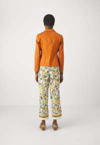 Giacca aderente arancione abbinata a pantaloni dai motivi floreali colorati con foglie verdi, rosa e gialle. Scarpe con zeppa in legno visibili.