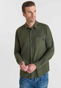 Man in een olijfgroene knoopshirt met borstzak en blauwe jeans, kijkt opzij, met zijn handen voor zich gekruist.