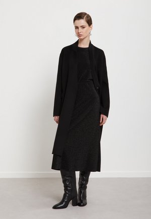 Donna che indossa un lungo cappotto nero, un vestito midi nero scintillante, stivali neri al ginocchio e orecchini a cerchio color argento su uno sfondo semplice.