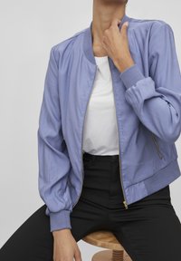 Ljuslila bomberjacka med ribbade manschetter, framdragkedja och avslappnad passform, i kombination med en vit t-shirt och svarta byxor.