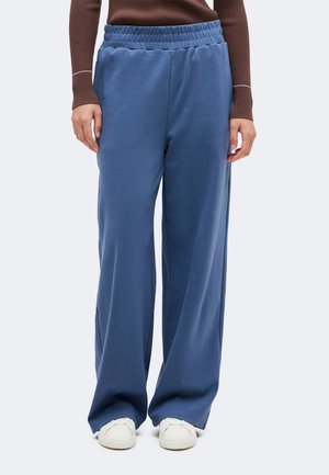 Modèle portant un pantalon bleu ample à jambes larges et à taille élastiquée, un haut marron et des baskets blanches.