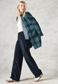 Zeleno-modrý kostkovaný oversize flanelový svetr, přeložený přes světle šedé tričko, doplněný tmavými širokými kalhotami a bílými teniskami.