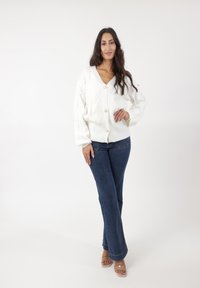 Hvid strikket cardigan med v-hals, prydet med et tekstureret mønster og runde knapper, parret med blå flagrende jeans og klare sandaler.