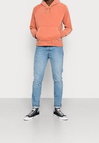 Felpa con cappuccio color corallo con lacci, tasca anteriore, abbinata a jeans azzurri chiari e sneakers nere alte. Tessuto liscio, vestibilità casual.