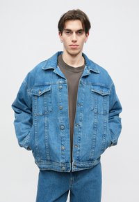 Τζάκετ από τζιν σε ανοιχτό μπλε χρώμα με χαλαρή γραμμή, εμπρόσθιο κλείσιμο με κουμπιά, δύο θήκες στο στήθος και εμφανείς ραφές. Φορώντας το πάνω από ένα γκρι πουκαμίσα.