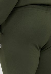 Olivgrüne Leggings aus dehnbarem Baumwollmaterial, mit glatter Textur und einem kleinen Logodetail an der Seite.