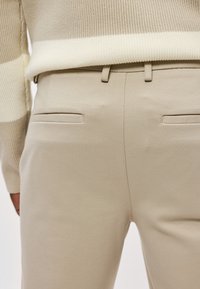 Beige räätälöidyt housut, joissa on sileä kangas, kaksi takataskua ja vyölenkit. Yhdistettynä kerma-valkopäällyksiseen neulepaitaan.