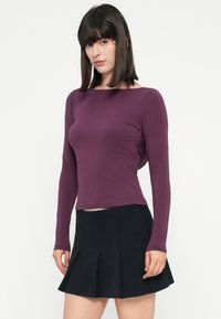 Donna con capelli lisci e scuri, che indossa un top attillato a maniche lunghe viola e una mini gonna nera plissettata, su uno sfondo bianco.
