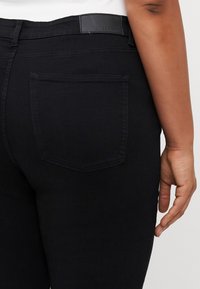 Jean en denim noir avec une coupe ajustée. Les caractéristiques incluent une poche arrière, une étiquette en cuir au niveau de la ceinture, et une surface lisse légèrement texturée.