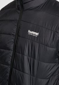 Doudoune noire avec des panneaux matelassés horizontaux, dotée d'un col haut, d'une fermeture zippée et d'un logo "hummel sportswear" blanc sur la poitrine.