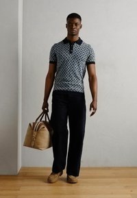 Granatowa koszulka polo w wzory, z krótkim rękawem, czarny kołnierzyk; ciemne spodnie; trzyma brązową torbę sportową z jasnobrązowymi akcentami; beżowe buty.