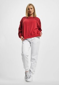 Rotes Sweatshirt mit strukturierten Ärmeln, lockerer Passform und abgerundetem Saum. Kombiniert mit weißen Sweatpants und weißen Sneakers mit schwarzen Akzenten.