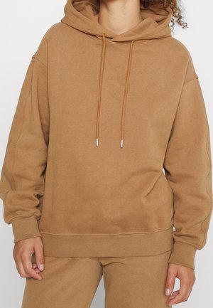 Bruine oversized hoodie met trekkoorden, gedragen met bijpassende bruine broek, waarbij de torso en handen zichtbaar zijn tegen een witte achtergrond.