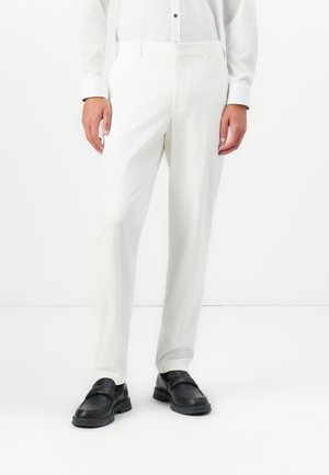 Man wearing white tailored trousers and black loafers, standing against a plain white background.