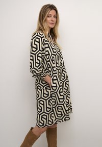 Vestido de patrón geométrico en negro y beige con un corte relajado, mangas largas, cierre de botones y bolsillos laterales. Combinado con botas marrones hasta la rodilla.
