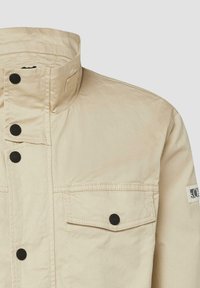 Bēša jaka ar augstu apkakli, melniem piespraužamiem pogām, krūšu aizslietņa kabatu un nelielu logotipa ielīmējumu uz augšējās piedurknes.