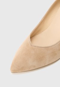 Escarpin beige en daim à bout pointu avec un design ajouré, une doublure intérieure lisse et des coutures discrètes le long du bord. Talon plat, style minimaliste.