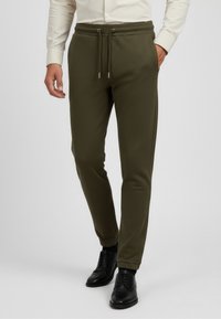 Olivgrön joggerbyxor i mjukt material, med midja som kan justeras med snöre, sidofickor och tapered ben. Svarta skor bärs för kontrast.