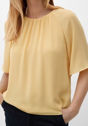 Blouse jaune, ample avec des manches courtes, col rond plissé et tissu doux et lisse. Des poches latérales sont en tissu plus foncé.