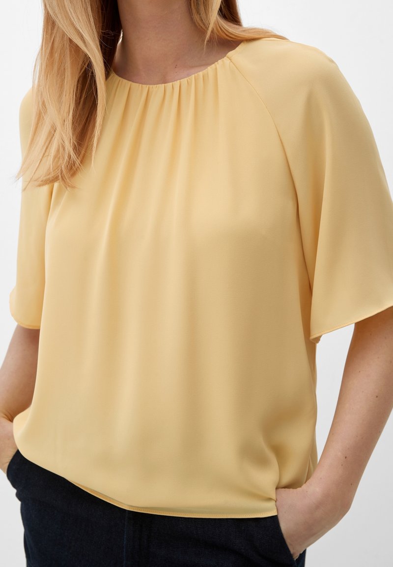 Blouse jaune, ample avec des manches courtes, col rond plissé et tissu doux et lisse. Des poches latérales sont en tissu plus foncé.