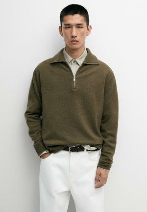 Giovane uomo che indossa un maglione verde oliva con zip sopra una camicia beige con colletto, pantaloni bianchi e una cintura intrecciata scura su uno sfondo semplice.