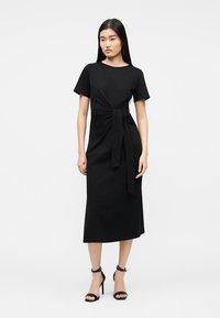 Γυναίκα στέκεται φορώντας μαύρο μίντι φόρεμα με κοντά μανίκια, ζώνη στη μέση, και μαύρα ψηλοτάκουνα πέδιλα με λουράκι στον αστράγαλο.