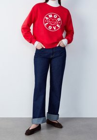 Sudadera roja con estampado en blanco de "AMOR LOVE", combinada con jeans de tiro bajo azul oscuro y zapatos planos marrones. Tejido ligero, ajuste casual.