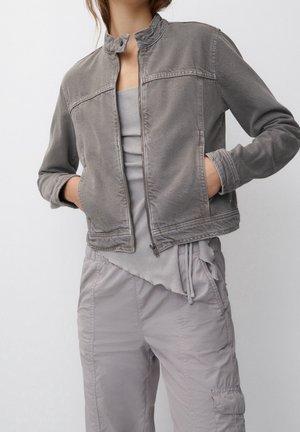 Mujer con chaqueta gris descolorida y sin cerrar sobre una camiseta de tirantes con canalé y pantalones cargo grises y holgados, manos en los bolsillos de la chaqueta, sobre un fondo liso.