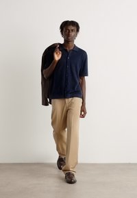 Camicia blu navy a coste con bottoni, pantaloni beige e scarpe marroni. Il modello tiene una giacca scura, in piedi su un pavimento chiaro davanti a uno sfondo semplice.