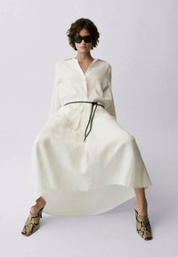 Robe blanche à manches longues, cintrée par une ceinture noire, avec une silhouette fluide. Accentuée par des mules à imprimé serpent et des lunettes de soleil sombres.