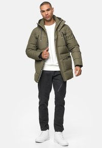 Olivengrøn pufferjakke med hætte, lynlås foran, to brystlommer og en tekstureret overflade, parret med sorte jeans og hvide sneakers.