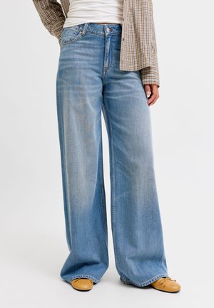 Personne portant un jean large bleu clair, des ballerines couleur sable, un top blanc et une chemise à carreaux marron, une main le long du corps.