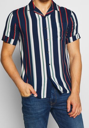 Muž v krátkém rukávu s košilí na knoflíky s vertikálními proužky v námořnické modři, bílé, zelené a červené barvě, v kombinaci s modrými džínami, s rukou v kapse.