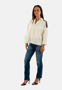 Camisa blanca de gran tamaño, con botones y rayas negras en los hombros, combinada con jeans ajustados azules y sandalias de tacón negras.