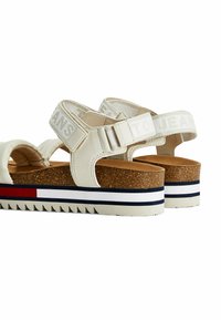 Sandali in pelle bianca con suola in sughero, caratterizzati da cinghie regolabili e una suola intermedia a strisce in rosso, bianco e blu navy. Il testo sulle cinghie riporta "PER I JEANS."