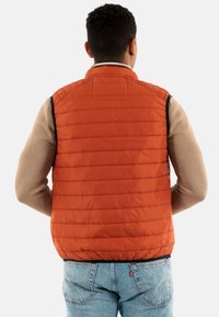 Gilet rembourré orange avec des coutures verticales, col montant et finitions en marine, porté par-dessus une chemise beige à manches longues, associé à un jean bleu.