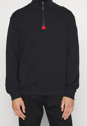 Man draagt een zwarte lange mouw pullover met een kwart ritssluiting en een rode ritstrekker, gecombineerd met een zwarte broek.