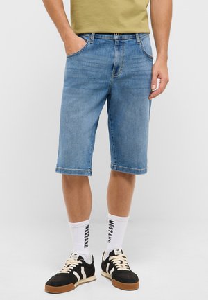 Man wearing light blue denim shorts, white branded socks, black and white sneakers, and an olive green shirt, standing with one hand in pocket.